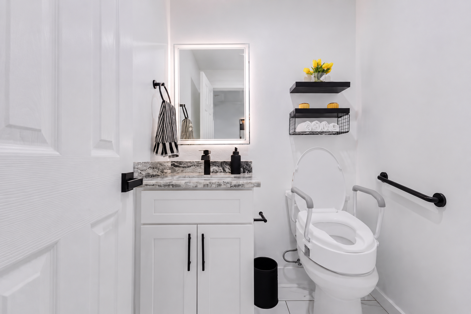 Accessible powder room with a raised toilet seat, grab bars, lit mirror and fresh towels